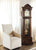 Reloj de pie de madera junto a un sillón, con frente de cristal y mecanismo visible