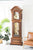 Reloj de pie en caja de madera marrón con frente repujado, ventana de cristal y pesas visibles.