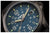 Primer plano de reloj de pulsera: esfera azul oscuro, números e índices luminosos, caja negra
