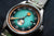 Reloj de hombre en primer plano: esfera turquesa, marcadores naranjas, correa de cuero, corona