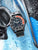 Reloj con esfera negra, bisel rojo, correa negra y corona lateral
