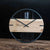 Reloj de pared redondo con esfera de madera clara, marco negro y manecillas de flecha.