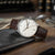 Reloj de hombre sobre mesa: correa de cuero marrón, esfera blanca con índices y logo