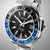 Reloj de hombre en primer plano: esfera negra, bisel azul, índices plateados