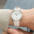 Reloj de mujer en la muñeca: correa de cuero beige, caja plateada, esfera blanca, manecillas azules