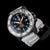 Reloj de hombre con esfera oscura, manecillas naranjas y correa de silicona blanca.