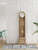 Reloj de pie de madera clara con esfera redonda, frontal de cristal y pomo blanco