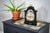Reloj de mesa negro con indicador de fase lunar dorado sobre base junto a planta en maceta