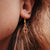 Colgante de oreja dorado con piedra oscura en marco cuadrado, visible en la oreja.