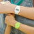 "Relojes digitales con correa blanca y verde en la muñeca, pantallas visibles"