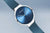 Pulsera de reloj en vista de 3/4: esfera azul, manecillas delgadas, correa milanesa