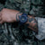 Reloj de hombre en muñeca con ropa de camuflaje: esfera azul, correa oscura, marcadores luminosos visibles.