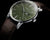 Reloj unisex con caja redonda, esfera verde y correa de cuero marrón sobre fondo oscuro.