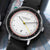 Reloj de hombre en vista oblicua con correa de cuero negra, esfera blanca y marcadores de minutos rojos.