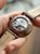 Fondo de reloj para hombre con movimiento expuesto en caja gris, correa de cuero marrón.