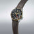 Reloj de hombre en vista lateral: correa de cuero marrón, esfera negra, bisel dorado.
