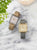 "Dos relojes de mujer estilo 'tank' con esfera blanca, caja dorada y correas de piel (gris/beige)."