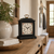 Reloj de mesa negro sobre superficie de madera, esfera blanca con marco floral, asa dorada