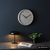 Runde Wanduhr mit hellem Zifferblatt und schwarzen Zeigern an dunkler Wand.