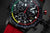 Reloj de hombre, vista de cerca: esfera negra con detalles rojos y pulsadores de cronógrafo