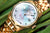 Reloj de mujer con bisel dorado, esfera azul e índices, visualización de fecha a las 5 en punto.