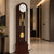 Reloj de pie con caja de madera oscura, esfera blanca y péndulo dorado en cristal