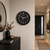 Reloj de pared redondo con esfera negra, manecillas y números en oro rosa, Hermle