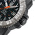 Detalle de reloj para hombre: caja estriada con punta de aguja naranja y protección lateral contra dobleces.