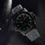 Reloj de hombre con caja y correa negras, índices luminosos en verde y naranja, bisel angular.