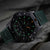 Reloj de hombre con caja negra, correa verde e índices luminosos en la esfera.