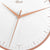 Primer plano de reloj de pared: aro metálico color rosa, esfera blanca y manecillas finas